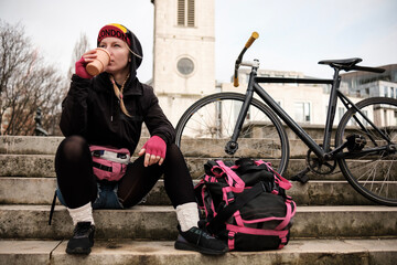 Portrait of bike courier woman taking a break and drinking coffee on stairs.