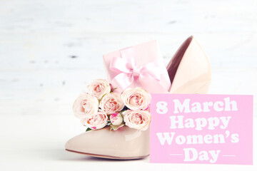 Bouquet of roses, gift box and card with text 8 March Happy Womens Day, and beige high-heeled shoe on white wooden background