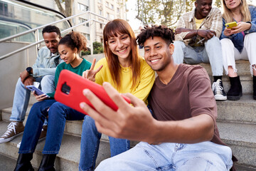 Multiracial young friends sitting on stairs hanging out, using phone to take photos and send...