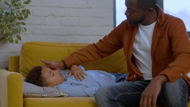 Seasonal virus. Little sick african american boy napping on sofa, caring father checking body temperature, empty space
