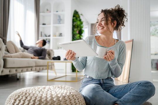 Young woman caucasian female sitting at home with cup of coffee use digital tablet while her boyfriend or husband is resting in background on the sofa bed real people leisure family concept copy space