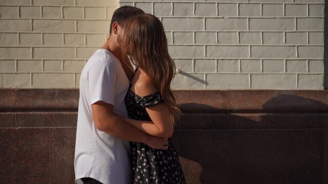 Loving hugging couple kissing in slow motion standing on city street outdoors. Side view medium shot portrait of confident tender Caucasian boyfriend and girlfriend dating enjoying leisure together