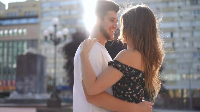 Side view zoom in to happy hugging couple talking standing on city street. Smiling Caucasian millennial boyfriend and girlfriend embracing in sunbeam outdoors in slow motion