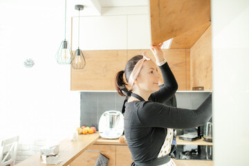 Happy woman open door of upper kitchen cupboard, side view. Housewife in headband and apron cook...