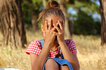 Little girl hiding face portrait. A kid in denim sundress sits in a field covering face with hands. A child playing hide-and-seek in summer meadow. Bullying, Abuse, and Child Protection Concept.