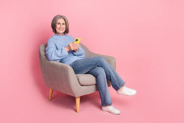 Full length photo of grey bob hair mature aged woman sit comfort chair wear blue sweater use phone for remote job isolated on pink color background
