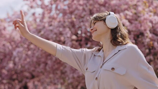 Carefree young woman walking and dancing in the blossom garden while listening music in wireless headphones. Beautiful girl enjoying leisure time outdoors using modern gadget