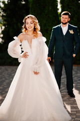a bride in a delicate dress stands in front of the groom in the park.