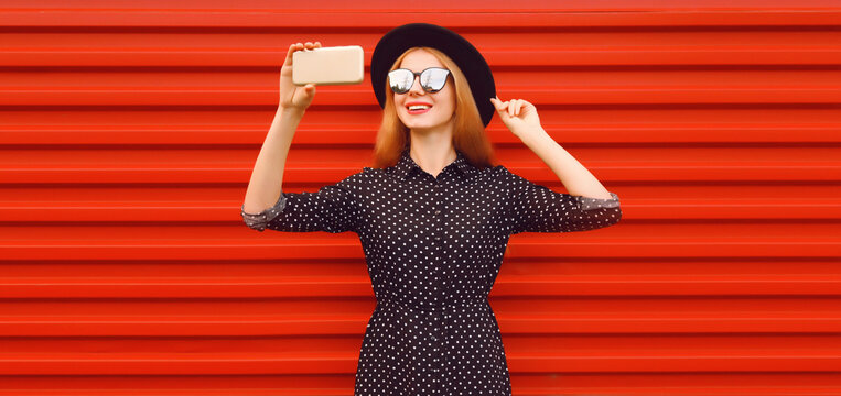 Portrait of happy smiling young woman taking selfie with smartphone wearing black round hat on red background