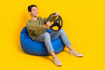 Full length photo of positive carefree man sit comfy bag hands hold wheel look empty space isolated on yellow color background