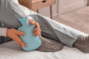 Young man warming his leg with hot water bottle in bedroom