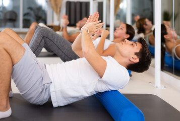 Slim young man practicing pilates with blue roller in training area during pilates classes