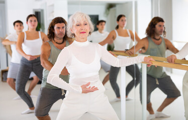 Fototapeta premium Gracile old-aged woman engaging in ballet at ballet barre in training hall during workout session