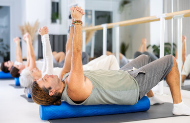 Fototapeta premium Diligent middle-aged man practicing pilates with blue roller in training area during pilates classes