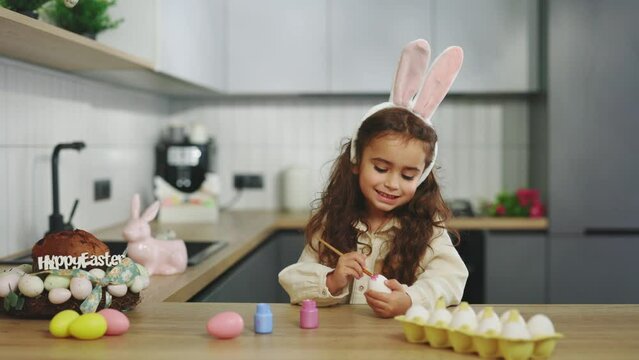 Beautiful little girl paint and decorate eggs for a traditional spring dinner at home. Little kid playing, drawing and decorating a pink colored egg. Happy easter
