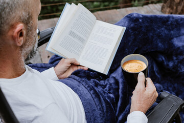 Hispanic man reading a book in the morning and holding coffee cup sitting down on the terrace of a...