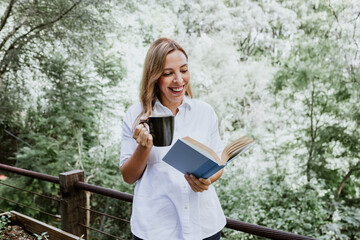Hispanic woman reading a book in a  morning and holding coffee cup on the terrace of a country...