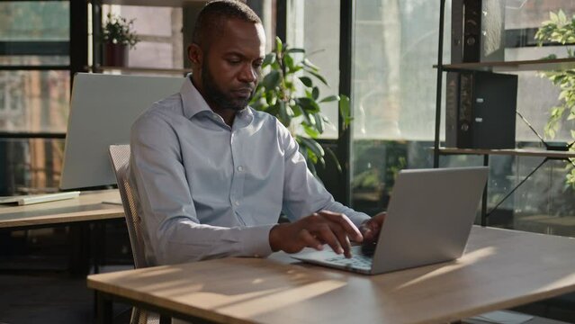 Focused adult senior businessman mature entrepreneur African American ethnic man working in office business firm programming on computer keyboard online project typing email sit in sunbeams background