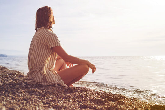 Young beautiful girl in a striped dress is resting on a pebble beach by the sea at sunset on a summer day, soft selective focus