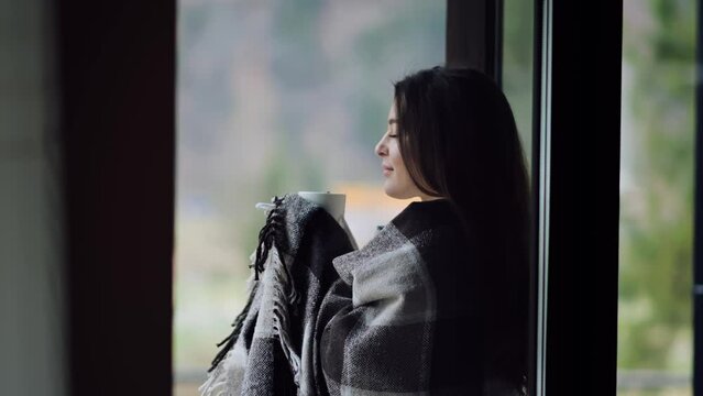 A woman with a cup of coffee or tea, a blanket on her shoulders, goes out to the balcony to admire the scenery