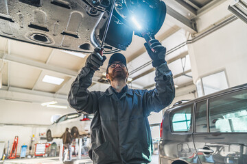 An expert mechanic with an LED light inspects a car on a lift in a repair station. High-quality...