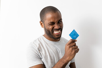 A curious dark-skinned man holds a condom while squinting his eye and looking at the camera,...