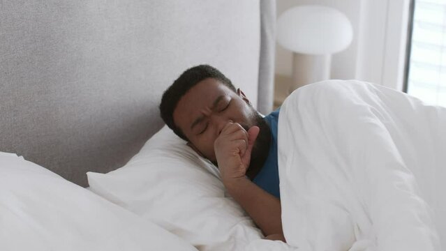 Young black african american man sleeping in bed but waking up with cough attack, keep resting at home in morning