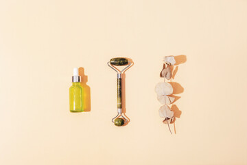 Green cosmetic serum bottle, jade facial roller and eucalyptus branch on neutral background with sharp shadows. Natural cosmetics, skin care concept. Top view, flat lay
