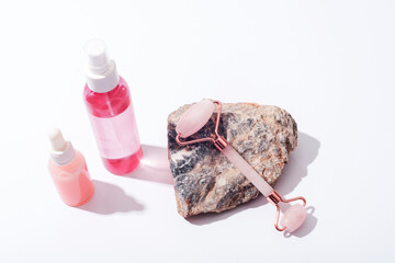 Pink cosmetic bottles and rose quartz crystal facial roller on natural stone on white background....