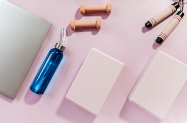 Set of accessories for fitness, yoga. Bra, pink block for yoga, bottle for water, dumbbells, sneakers, skipping rope, laptop on pink pastel background. Sports gear, minimalistic background. Close up