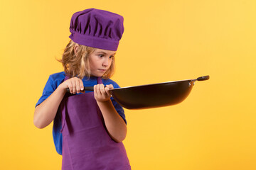 Kid cook with cooking pan. Child cooking, little chef prepares food. Kid boy in chefs hat and apron on yellow studio isolated background.