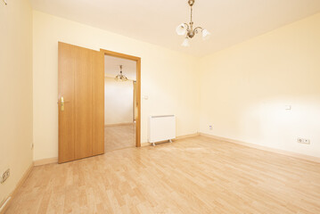 Fototapeta na wymiar Empty living room with light yellow walls, a light wooden floor, an electric heater, an oak wooden door and a ceiling lamp with a white shade