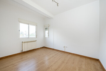 Empty living room with white walls, laminate flooring, white aluminum windows on two walls with blinds and a radiator below