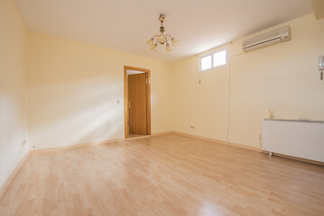 Empty living room with light yellow walls, light wooden floor, electric heater, oak wooden door, split air conditioner, skylights high on the walls and ceiling lamp with white shade