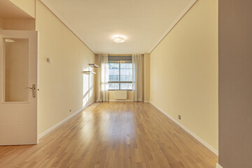 Empty living room with light yellow painted walls, white plaster moldings on the ceiling, aluminum windows, white curtains and an oak parquet floor