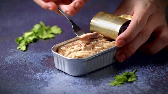 Hand with a fork picking up canned mackerel on a dark blue table with parsley. Ready to eat.