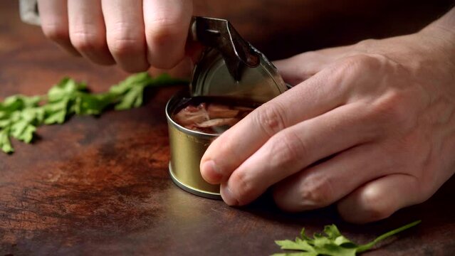 Hands opening canned octopus with a old can opener on a dark brown table with parsley. Ready to eat.