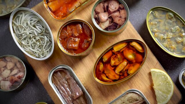 Preserve cans with different products, fish and seafood, natural or pickled, open on a dark table with a wooden cutting board and lemon. Ready to eat.