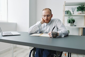 A man in a wheelchair, a businessman in the office works at a laptop, writing down a plan in a...