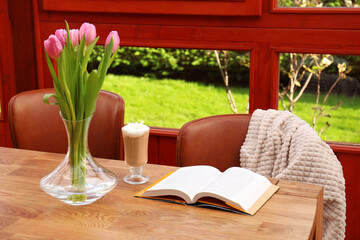 Glass of delicious cocoa, pink tulips and book on wooden table at terrace