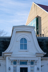 Obraz premium The exterior upward view of two buildings. A small wooden house with a bell shaped dormer and a closed arch shaped glass window. There's a large tan colored brick building with a red metal roof. 
