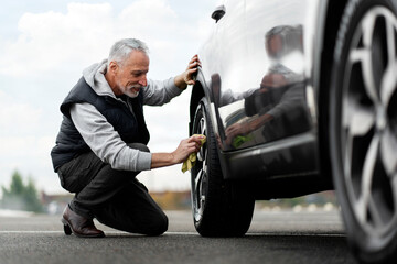 Happy senior man cleaning tyre and alloy wheel with microfiber while washing car at car wash station