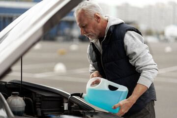 Gray-haired mature man driver filling the car's reservoir with the blue fluid - windshield washer