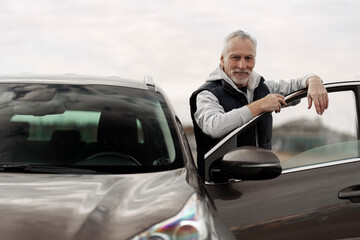 Handsome bearded senior man in dealership's parking lot, looking at camera, near his new automobile