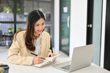 Asian businesswoman taking notes on her notebook, looking at laptop screen