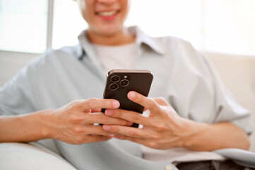 A happy young Asian man relaxing on sofa in his living room, using his phone, texting