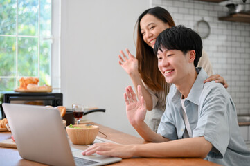 Happy young Asian couple saying hi to their parents through a video call on laptop in the kitchen.
