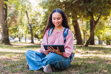 Latin university student woman smiling and using a tablet sitting outside in a park on the grass