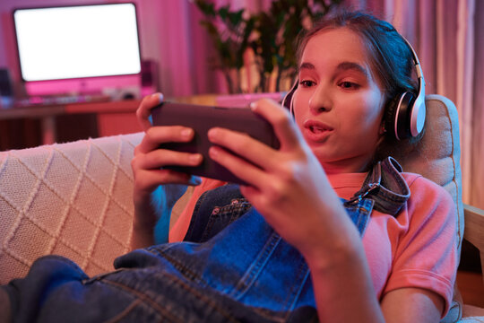 Excited teenage girl relaxing on sofa and watching videos of her favorite blooger on smartphone