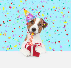 Jack russell terrier puppy wearing a party cap blows into party horn and holds gift box above empty white banner